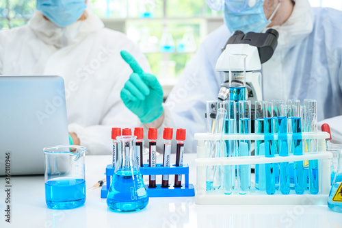 Lab equipment with blue liquid inside stand on the table with couple male scientist wearing protection suit at laboratory at the background.