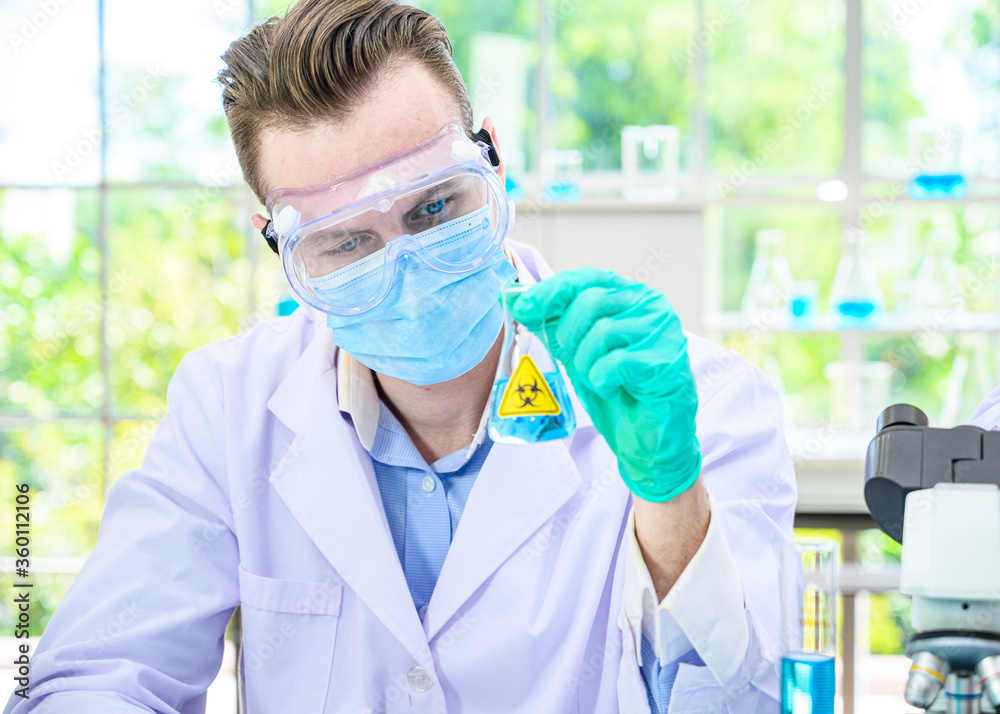 Male scientist wearing protection suit holding beaker or tube with sample liquid inside and working with many lab equipment for research at laboratory.