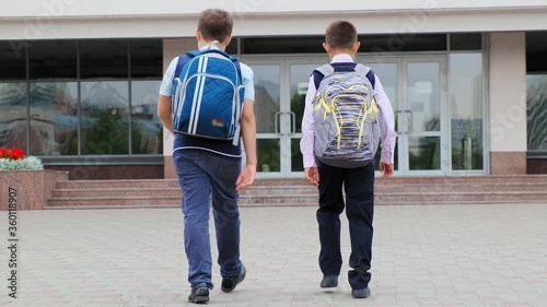 junior schoolboys with backpacks in uniform walk together to modern school building on summer day backside view slow motion