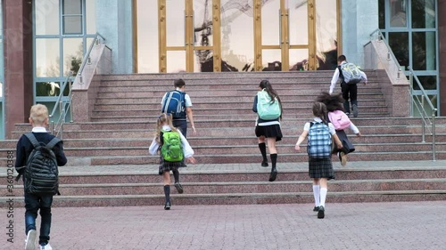 group of little pupils in uniform with schoolbags run up stairs to school building on warm day backside view slow motion