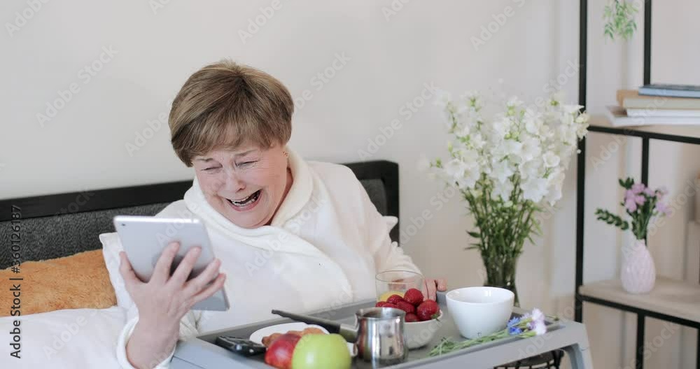 Old woman in home rob laughing while looking at screen of her modern device. Elderly female retiree having good mood while using tablet during breakfast in bed. Concept of emotions.