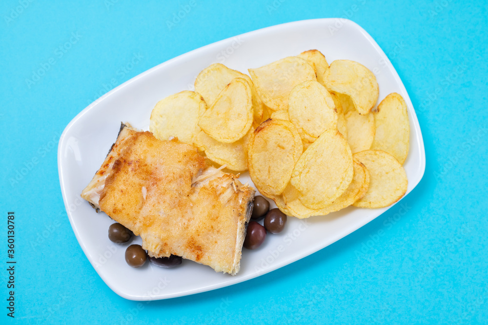 fried cod fish with olives and potato chips on white dish on blue background fried cod fish with olives and potato chips on white dish on blue background