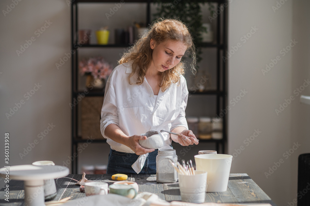 Female potter sitting and stirs paint with a brush a cup on the table. Woman making ceramic item. Pottery working, handmade and creative skills. Female potter sitting and stirs paint with a brush a cup on the table. Woman making ceramic item. Pottery working, handmade and creative skills.