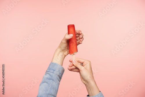 Horizontal shot of young female's hands with nude manicure posing over pink background with ice-cream on stick with red glaze. Food photography concept