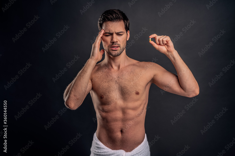 Handsome young bearded man isolated. Portrait of naked muscular man is standing on gray background and holding a pill. Man holding his temple. Experiencing headache.