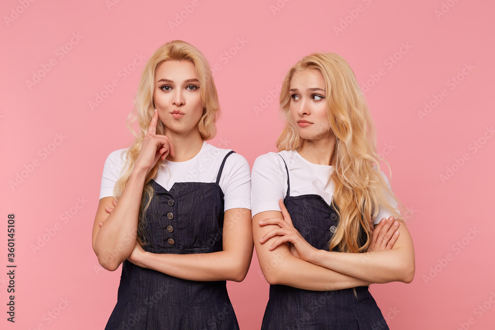Indoor photo of young brown-eyed long haired female keeping forefinger raised while looking attentively at camera, posing over pink background with her confused pretty blonde sister Indoor photo of young brown-eyed long haired female keeping forefinger raised while looking attentively at camera, posing over pink background with her confused pretty blonde sister