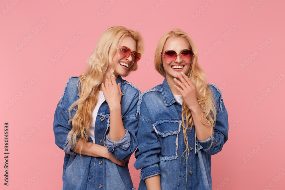 Indoor shot of young lovely happy long haired blonde twins dressed in same clothes having nice time together and laughing happily while standing over pink background