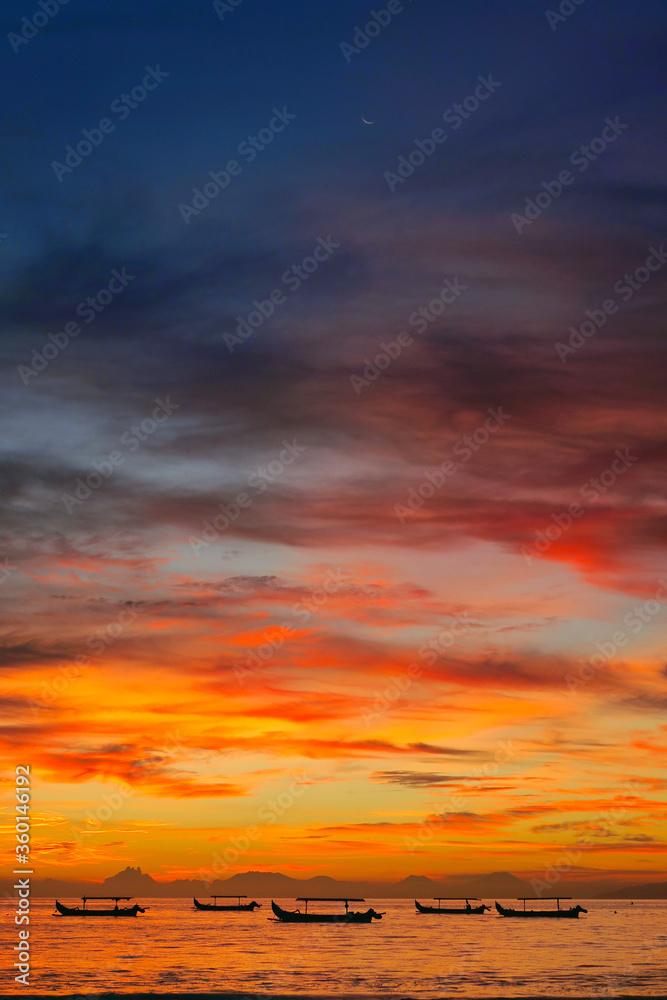 Fototapeta premium Fishermen boats silhouettes in tropical fiery sunset light at Kuta beach, Bali island, Indonesia. Vertical photo.