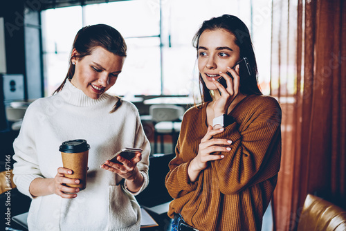 Successful hipster girl updating profile in social networks and installing app using 4g connection holding takeaway coffee while happy female friend making positive cellular call via application