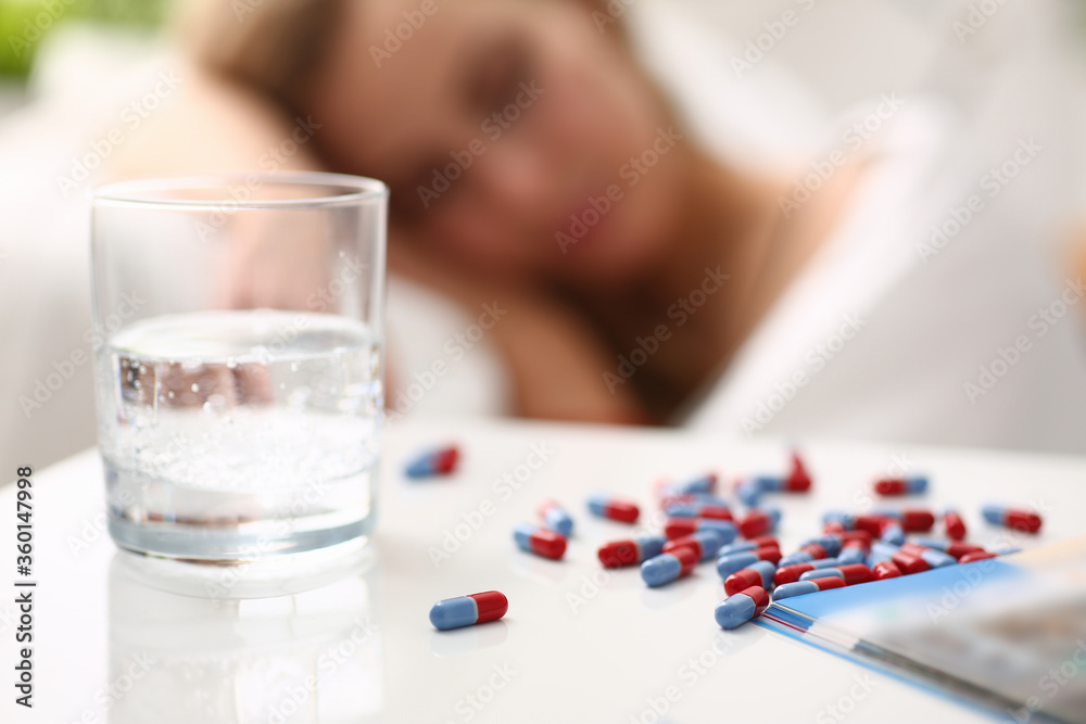 Close up of drugs and glass of water on white desk with sleeping lady on blurred background