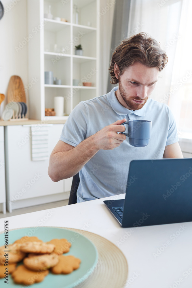 Young man sitting at the table working on laptop computer and drinking coffee in the morning in the kitchen Young man sitting at the table working on laptop computer and drinking coffee in the morning in the kitchen