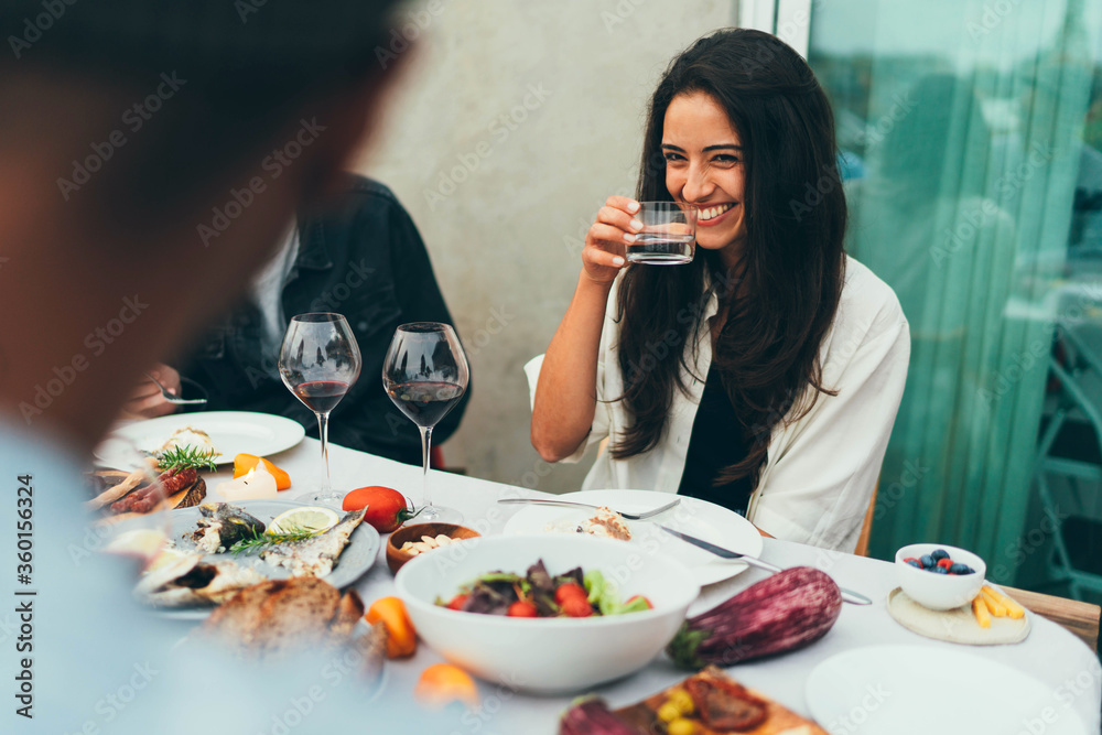 Attractive young woman with beautiful long hair laughing while drinking water at dinner party with friends, beautiful young girl playfully looks at the guy who sitting opposite her
