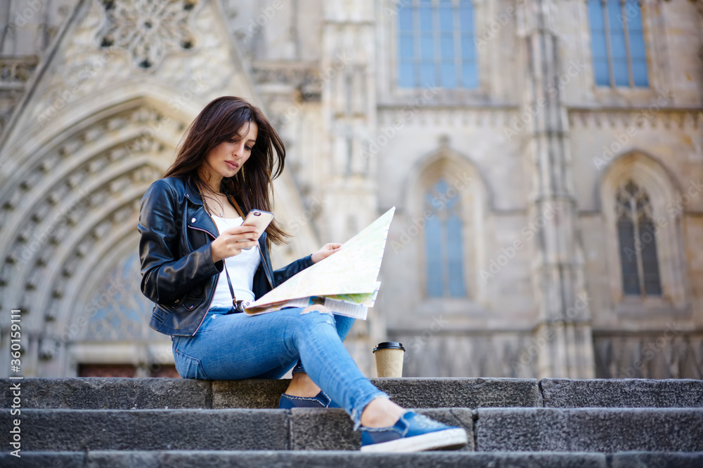 Charming brunette traveler using application on smartphone for searching destination, young woman in stylish clothes finding route on map and via navigation on mobile sitting on urban background