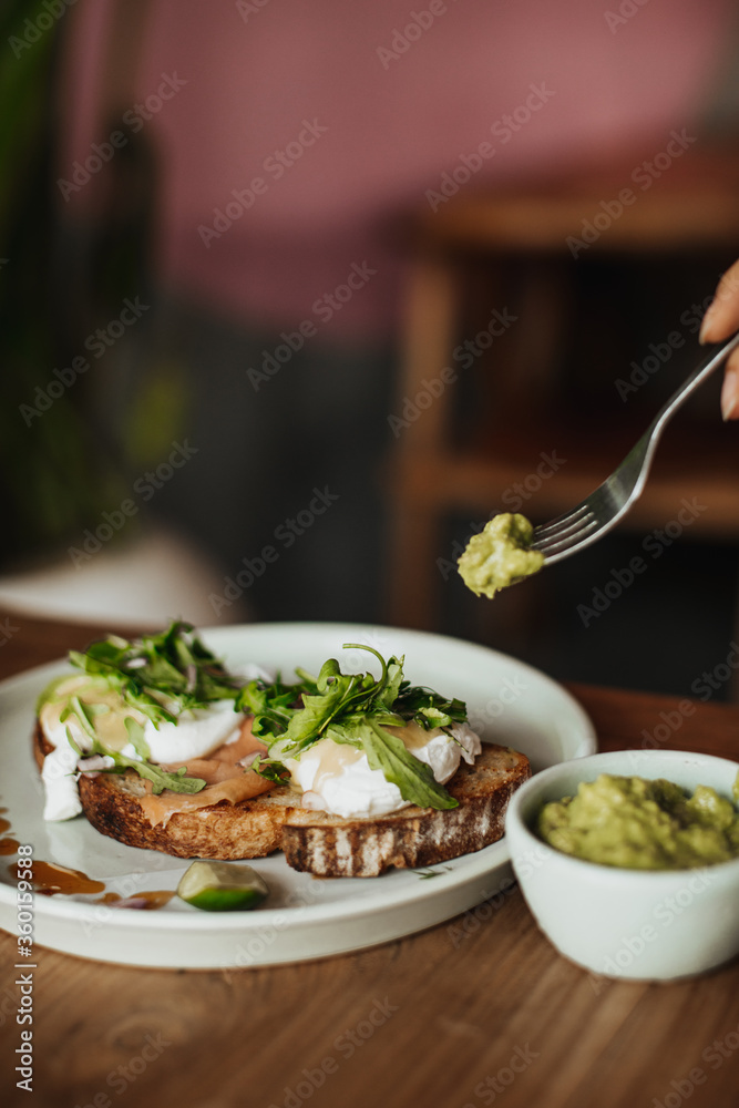 Photo of delicious guacamole sauce, plate with toast with salmon and poached eggs on white plate on wooden table