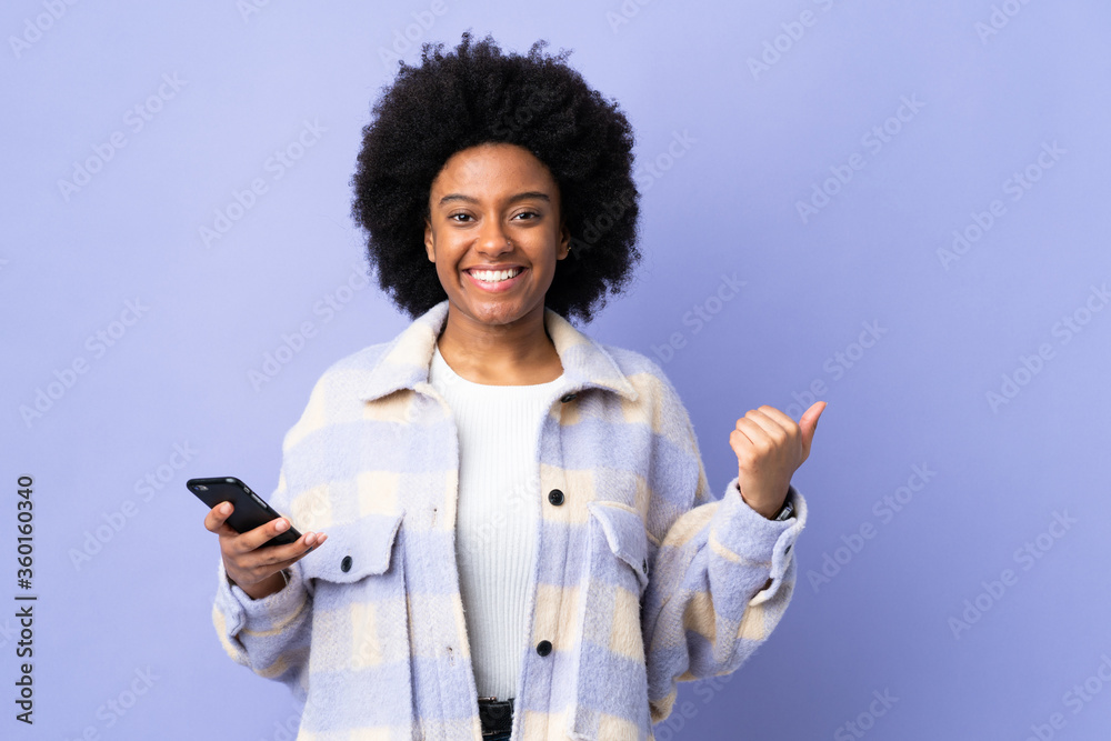 Young African American woman using mobile phone isolated on purple background pointing to the side to present a product Young African American woman using mobile phone isolated on purple background pointing to the side to present a product