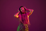 Young caucasian woman dancing with headphones on pink studio background in neon light. Beautiful model with dreadlocks. Human emotions, facial expression, sales, ad concept. Freak's culture.