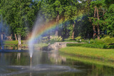 a pond with a fountain in a city park and a rainbow is visible through a stream of water