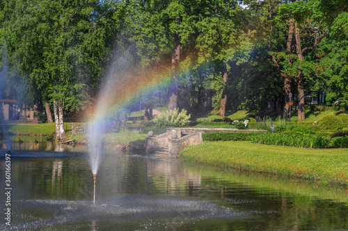 a pond with a fountain in a city park and a rainbow is visible through a stream of water