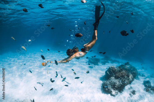 Freediver young woman with fins glides over sandy bottom with tropical fishes in transparent ocean