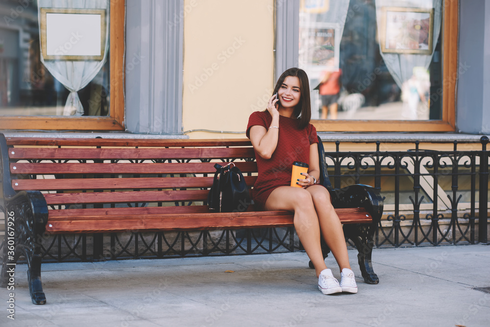 Happy woman having mobile phone conversation via cell telephone, while is resting on bench with cup of take away coffee. Smiling female student talk via cell telephone, while is resting after lectures Happy woman having mobile phone conversation via cell telephone, while is resting on bench with cup of take away coffee. Smiling female student talk via cell telephone, while is resting after lectures