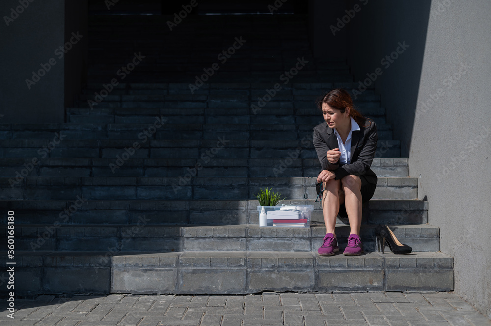 Depressed woman dressed in medical mask is fired and is sitting on the stairs with a box of personal belongings. Female office worker in suit and sneakers outdoors. Unemployment in the economic crisis Depressed woman dressed in medical mask is fired and is sitting on the stairs with a box of personal belongings. Female office worker in suit and sneakers outdoors. Unemployment in the economic crisis