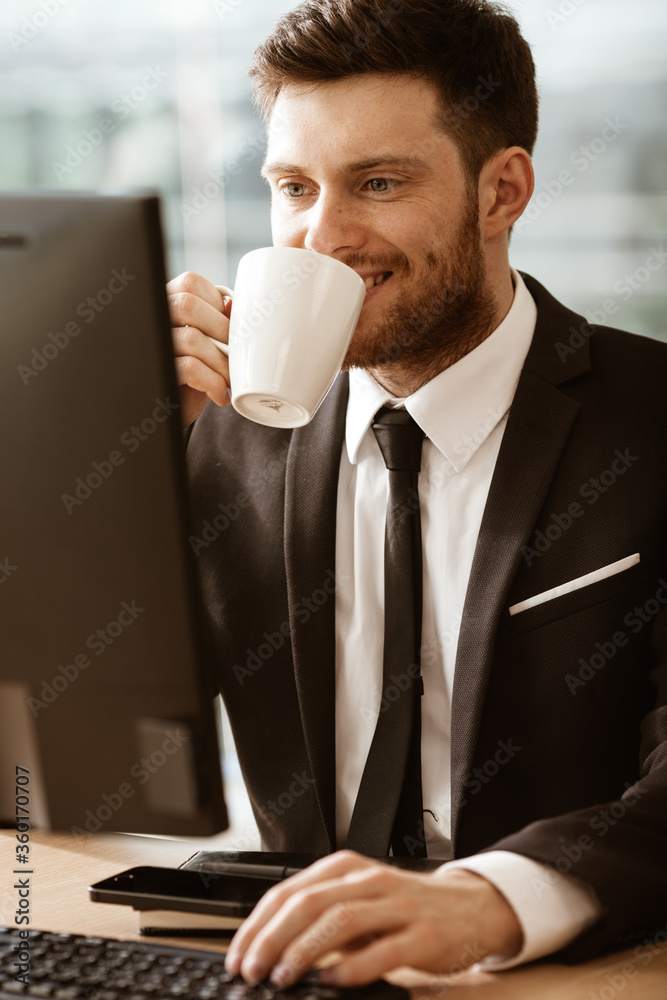 Business concept. Successful young businessman at work. Manager sitting at the office table happy drinking coffee from cup. Man smiling in suit indoors on glass window background Business concept. Successful young businessman at work. Manager sitting at the office table happy drinking coffee from cup. Man smiling in suit indoors on glass window background