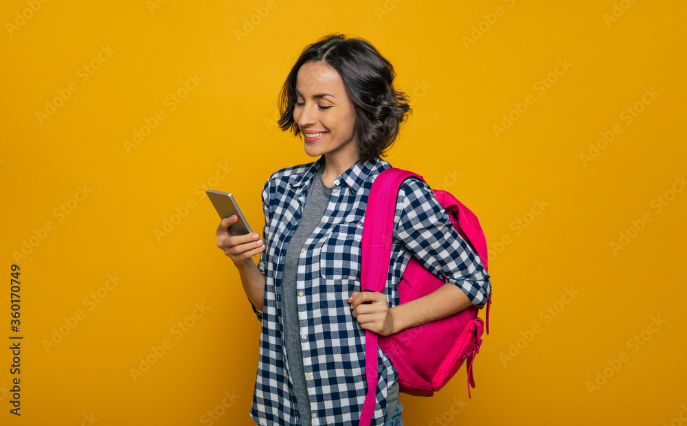  What's new on the internet? A half-length photo of a young beautiful student, dressed casually, with her cute pink backpack on her one shoulder, looking with a wide smile in her smartphone.