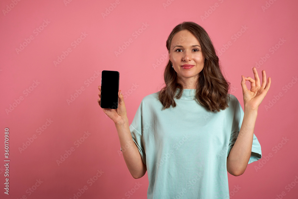 Beautiful girl holds mobile phone and makes okay gesture on a pink background. Emotions and pleasant feelings concept. Beautiful girl holds mobile phone and makes okay gesture on a pink background. Emotions and pleasant feelings concept.