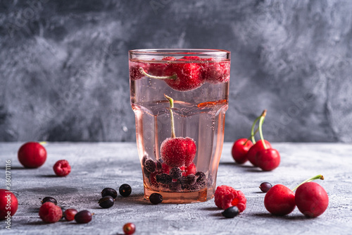 Fresh cold sparkling water drink with cherry, raspberry and currant berries in red faceted glass on stone concrete background, summer diet beverage, angle view