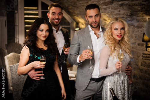 Party, holiday with friends concept. Four people with champagne glasses celebrating and toasting in restaurant. Two men and two women in elegant evening clothes, suits and dresses night out indoors