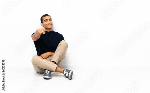 African American man sitting on the floor points finger at you with a confident expression
