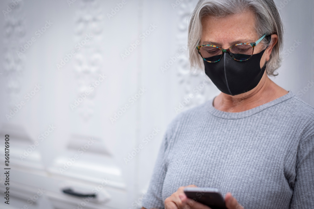 A white-haired senior woman with medical mask due to coronavirus standing against a white door using her mobile phone - relaxed retirement concept