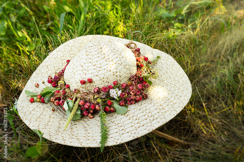 Wreath of wild berries on light straw hat on a green grass background. Fresh, ripe wild strawberry wreath, flowers and leaves on summer hat. Summer oundoor concept. photo