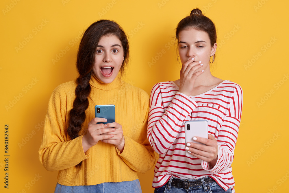 Two friends with phones reading shocking news in social network, lady with pigtail looking at camera with big eyes and yelling something, female in striped shirt covering her mouth with palm. Two friends with phones reading shocking news in social network, lady with pigtail looking at camera with big eyes and yelling something, female in striped shirt covering her mouth with palm.