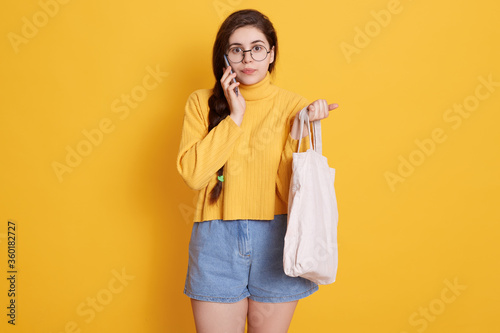 Adorable brunette woman wearing yellow sweater and short, holding bag in hand, talking to her friend via modern smart phone, standing isolated yellow wall.