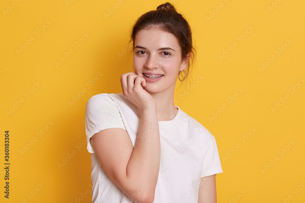 Adorable dark haired woman in brackets looking at camera with charming smile, holding her fist under chi, girl with hair bun dresses white casual t shirt. Adorable dark haired woman in brackets looking at camera with charming smile, holding her fist under chi, girl with hair bun dresses white casual t shirt.