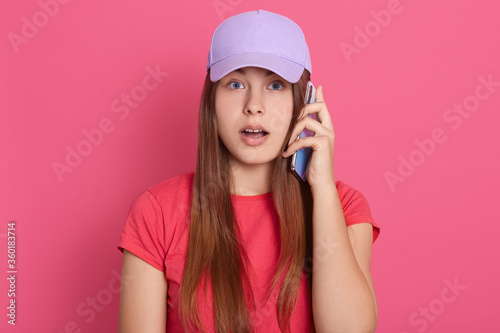 Lady in baseball cap and red t shirt talking via phone with astonished facial expression, female being shocked, hears breaking news, keeps mouth opened.