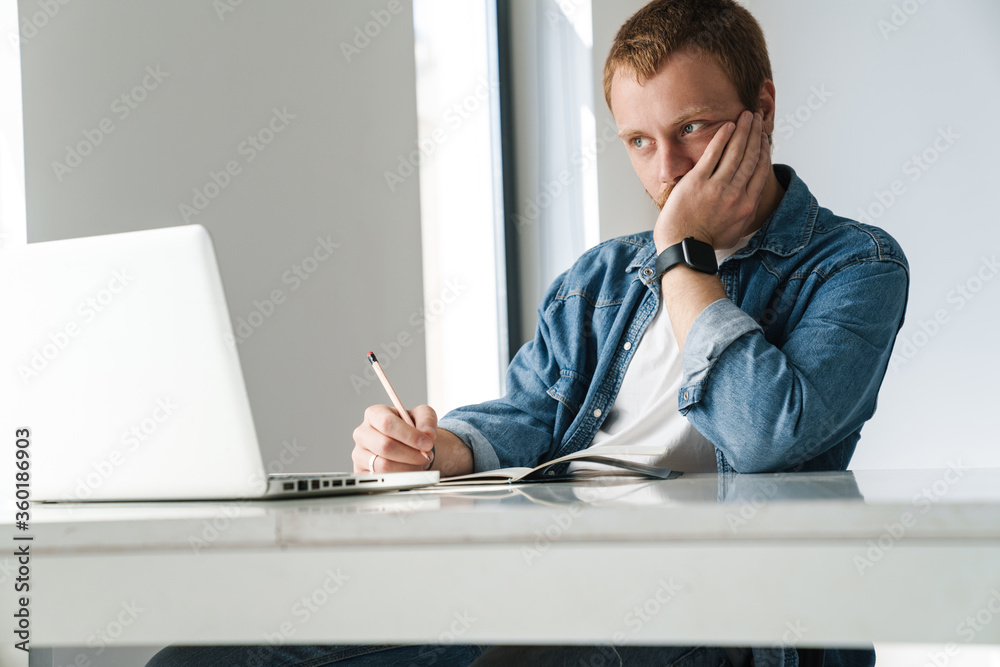 Photo of thinking man writing down notes while working with laptop Photo of thinking man writing down notes while working with laptop
