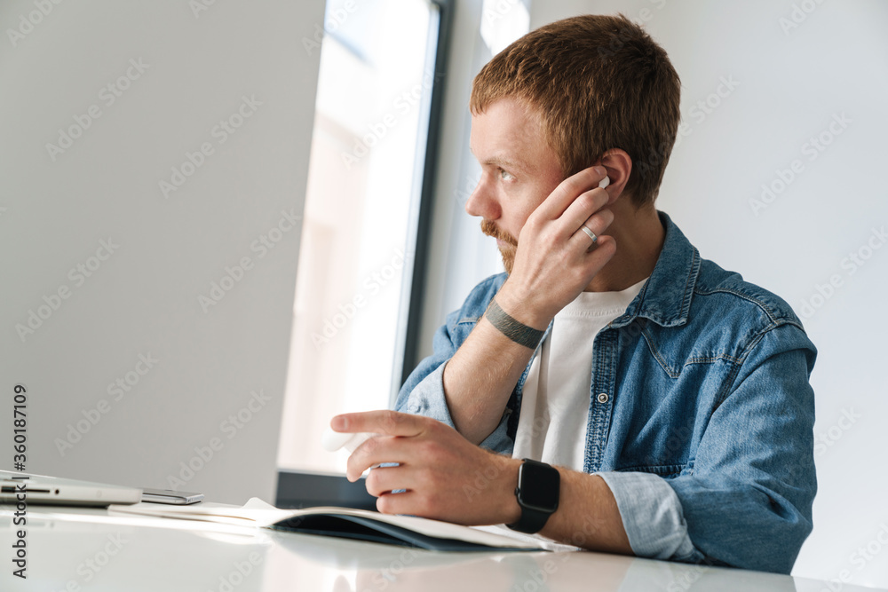 Photo of ginger man using wireless earphones while working with laptop