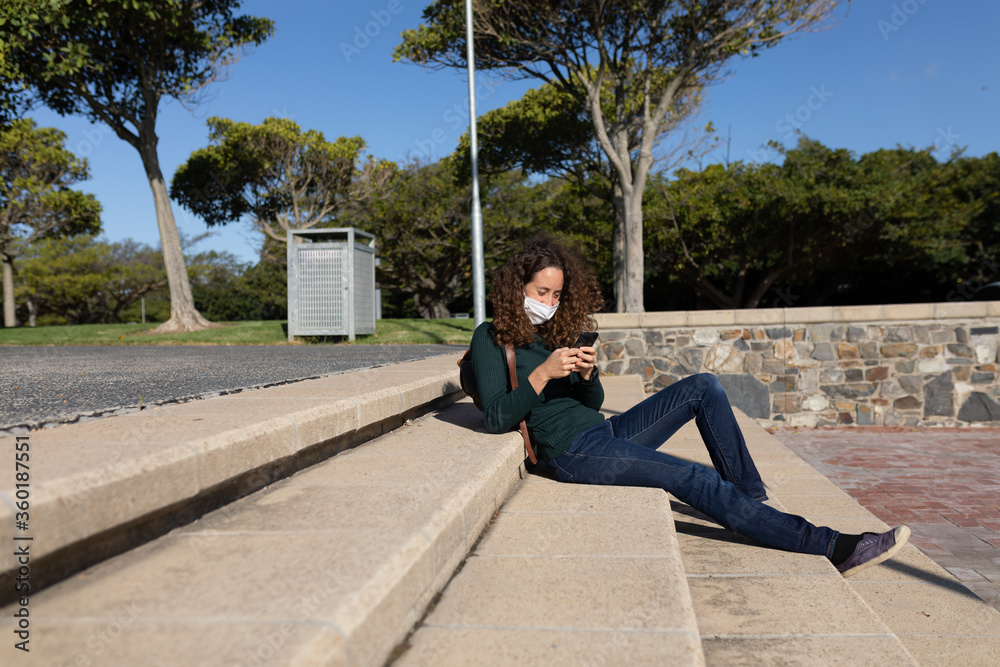 Caucasian woman wearing a protective mask out in the streets, sitting using her phone Caucasian woman wearing a protective mask out in the streets, sitting using her phone