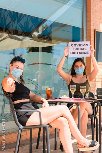 Two women wearing surgical masks and sitting at an outdoor terrace. Safety and social distancing concept.