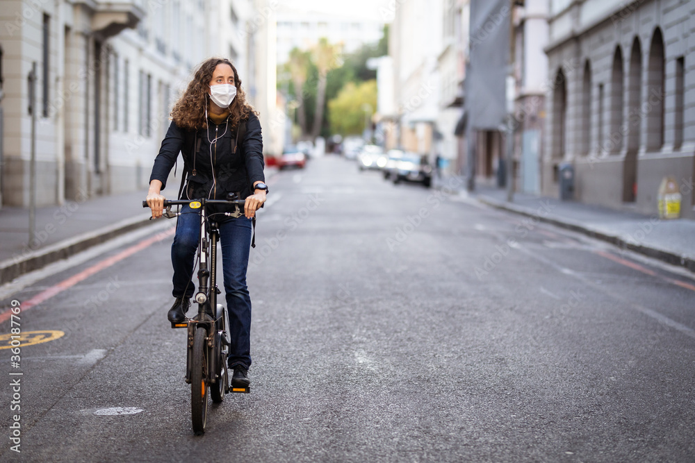 Caucasian woman wearing a protective mask and earphones, and biking in the streets