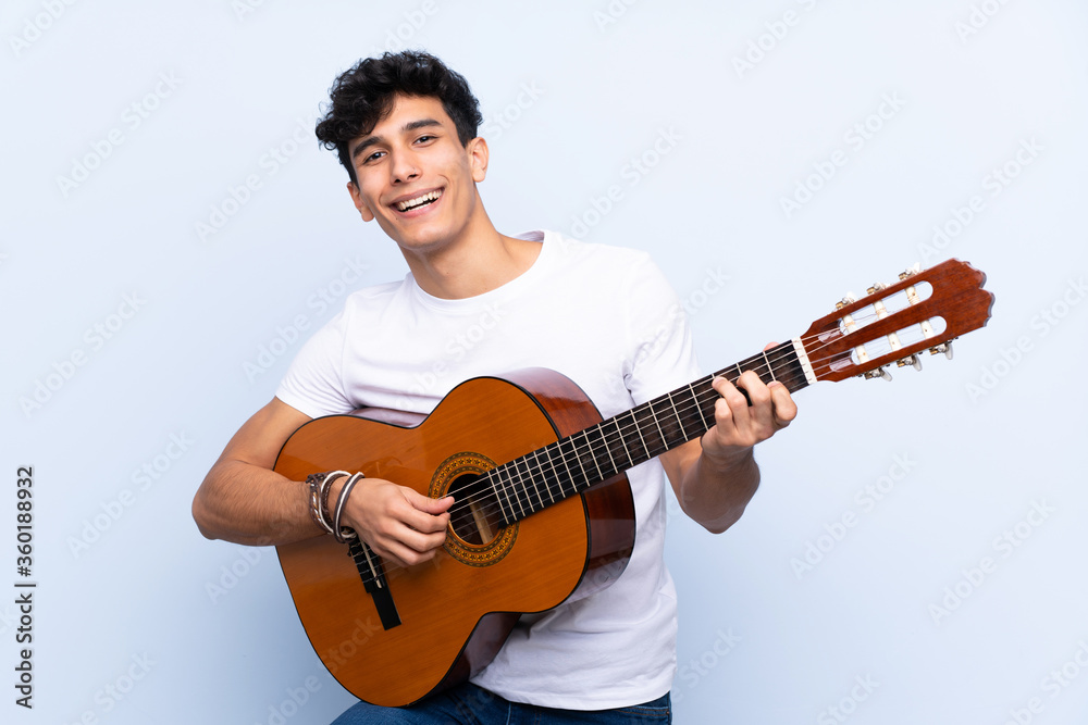 Young Argentinian man with guitar over isolated blue background