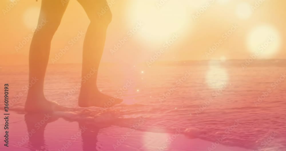 Digital composite video of low section of woman walking on the beach against glowing spots moving in