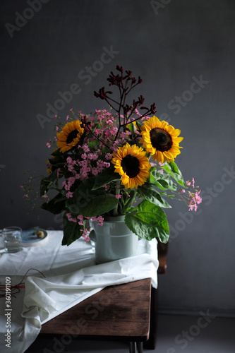 Üppiger Blumenstrauss mit gelben Sonnenblumen, Katzenpfötchen und kleinen lila Blumen auf einem Tisch vor einer grauen Wand bzw grauem Hintergrund