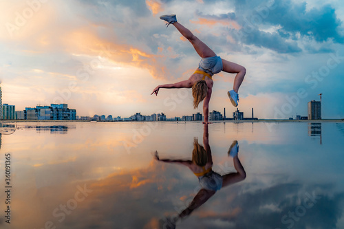 Photographie Female gymnast standing on one hand and keeping balance during dramatic sunset with reflection in the water of amazing clouds