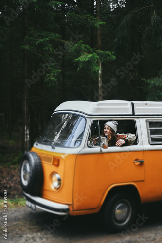 Stylish hipster girl sitting in orange retro Bulli Vintage Camper in green forest.