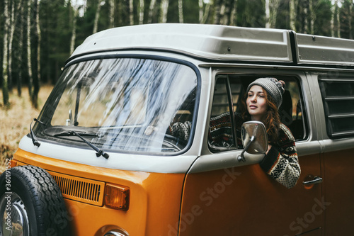 Stylish hipster girl sitting in orange retro Bulli Vintage Camper in green forest.