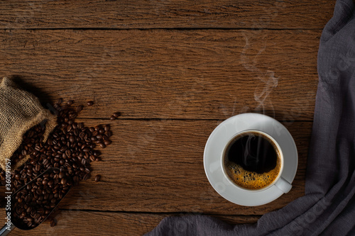 Top view above of Hot fresh black coffee with smoke and milk foam in a white ceramic cup with coffee beans roasted in sack bag on wooden table background. Flat lay with copy space.