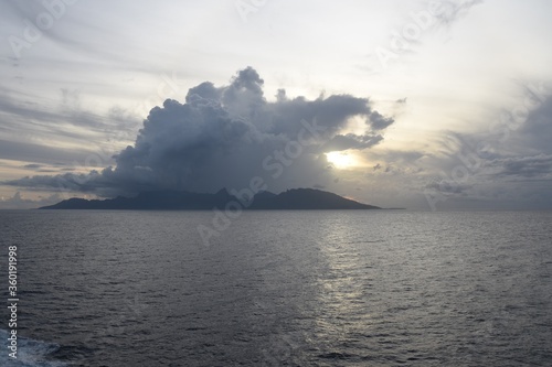 Island in French Polynesia, Pacific ocean during sunset with dramatic clouds and covered sun during calm weather and sea- Picture was taken from the ship